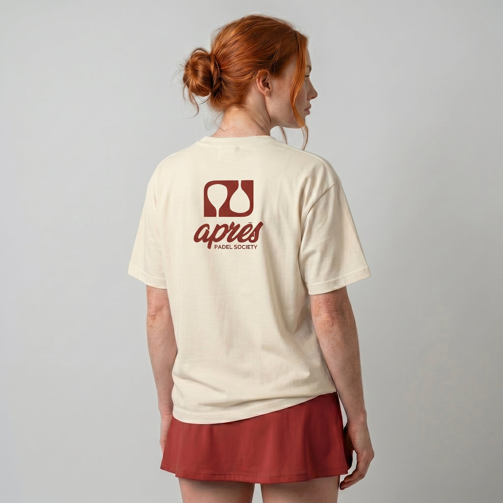 Female model with back toward camera wearing an oversized cream unisex padel t-shirt in relaxed fit with a red logo central on the back.