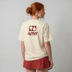 Female model with back toward camera wearing an oversized cream unisex padel t-shirt in relaxed fit with a red logo central on the back.