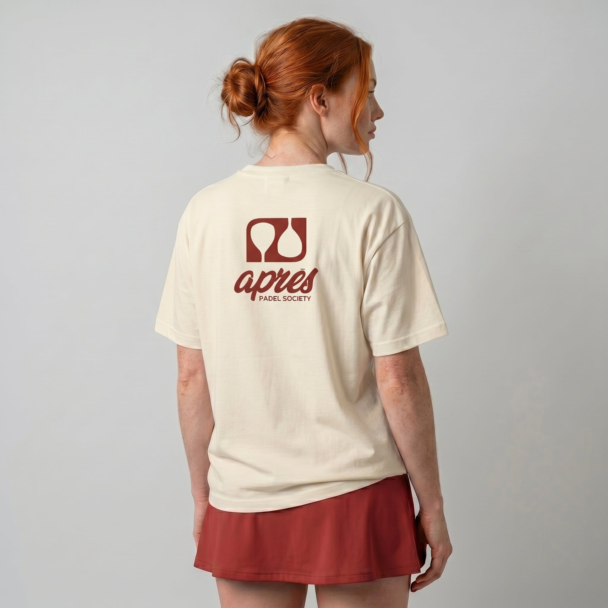 Female model with back toward camera wearing an oversized cream unisex padel t-shirt in relaxed fit with a red logo central on the back.
