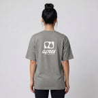 Female model with back toward camera wearing an oversized heather grey unisex padel t-shirt in relaxed fit with a white logo central on the back.