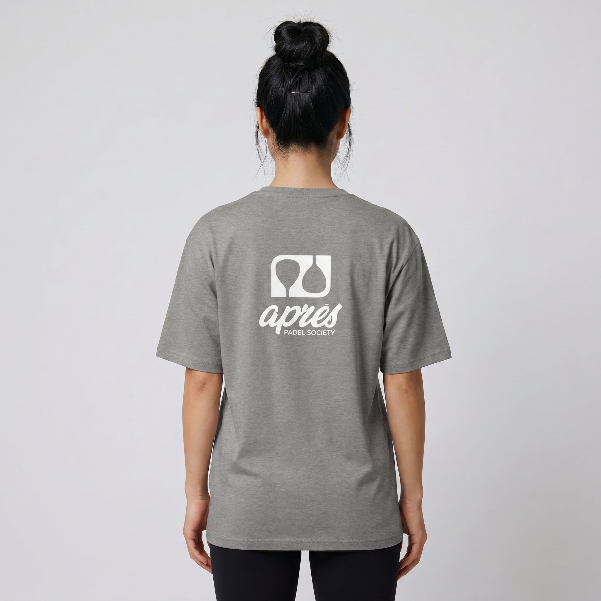 Female model with back toward camera wearing an oversized heather grey unisex padel t-shirt in relaxed fit with a white logo central on the back.