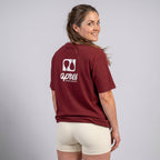 Female model with back toward camera wearing an oversized maroon unisex padel t-shirt in relaxed fit with a white logo central on the back.