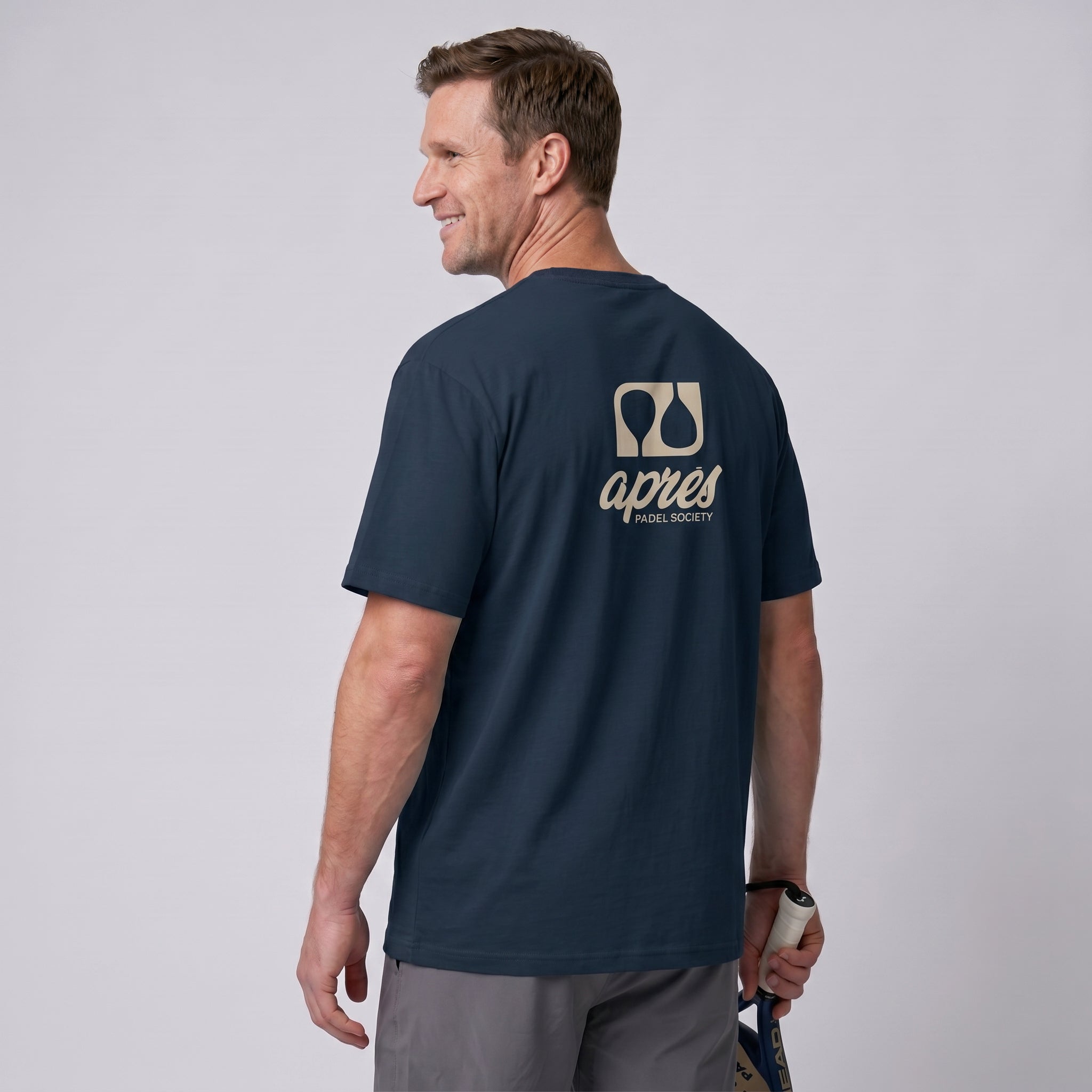 Male model with back toward camera wearing an oversized navy blue unisex padel t-shirt in relaxed fit with a tan logo central on the back.