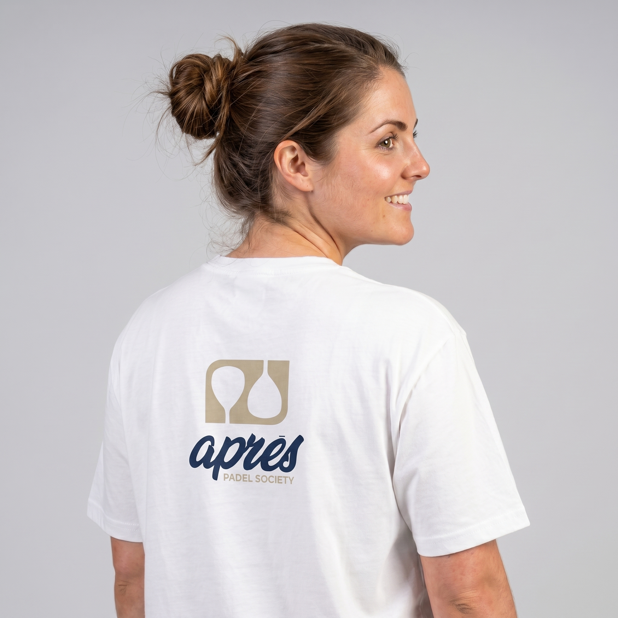 Female model with back toward camera wearing an oversized unisex white padel t-shirt in relaxed fit with a tan and navy logo central on the back.