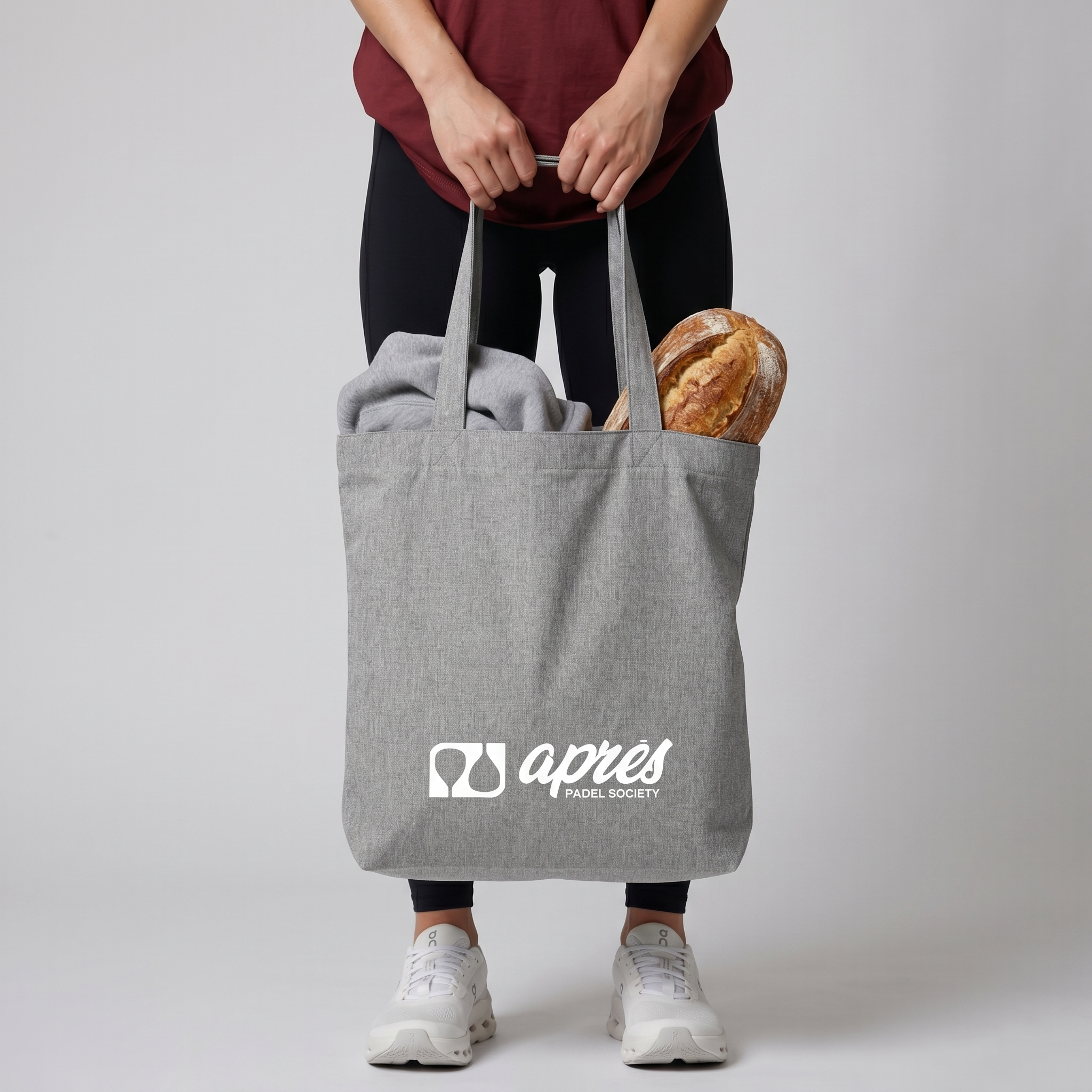 Model carrying a premium heather grey padel tote bag with visible hoodie and fresh bread inside, featuring a white 'Après Padel Society' logo.
