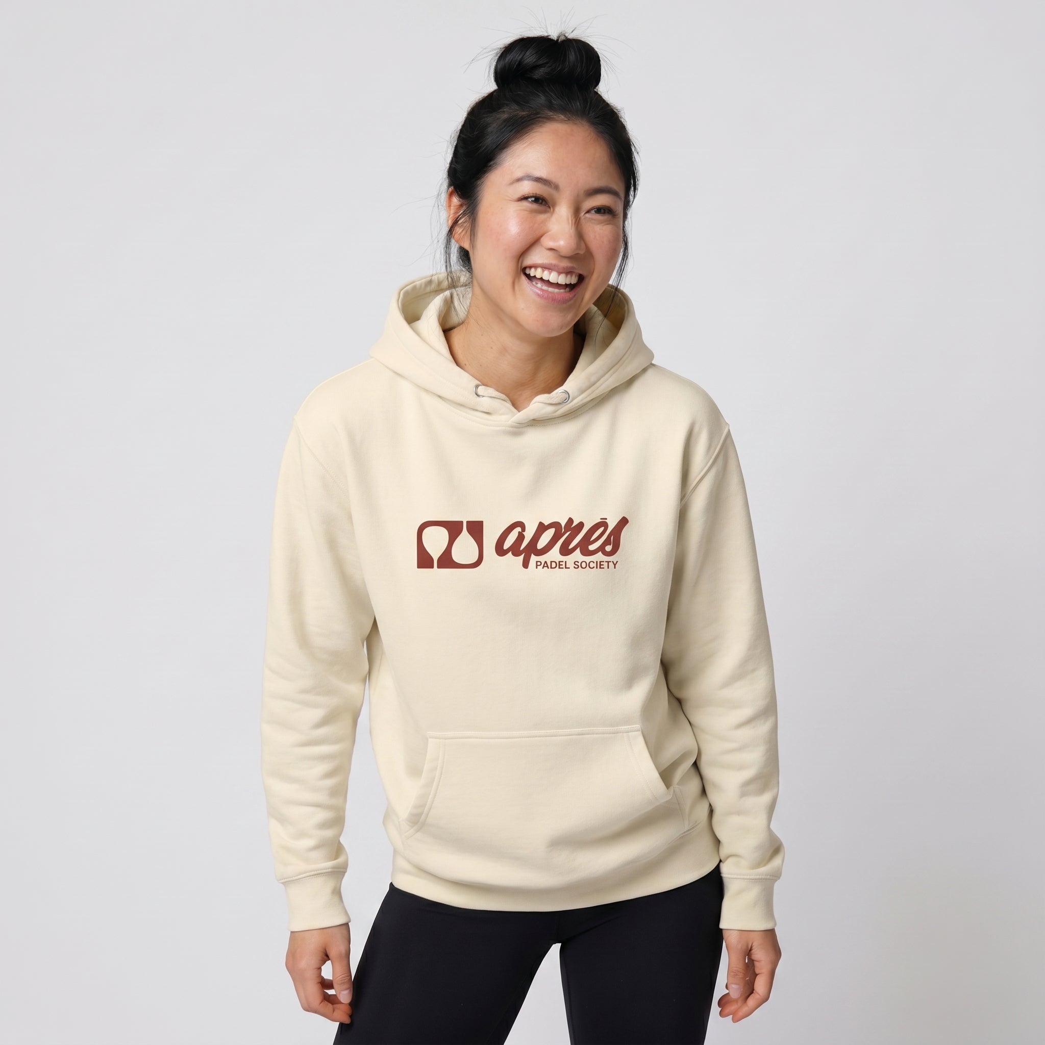 Woman modelling a cream premium padel hoodie with a red horizontal 'Après Padel Society' logo.
